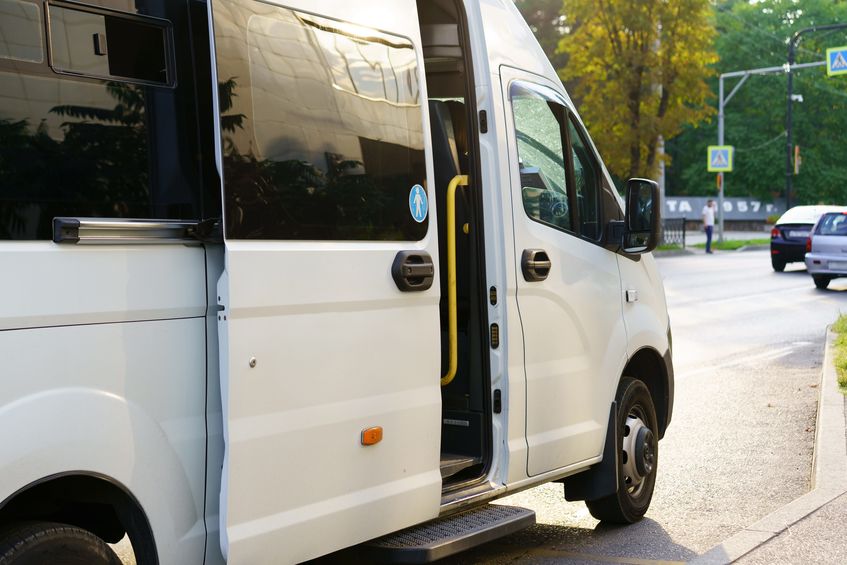 A white delivery van stands parked along a bustling city street in the afternoon. the doors are open, suggesting the driver is engaged in a drop-off or pickup.